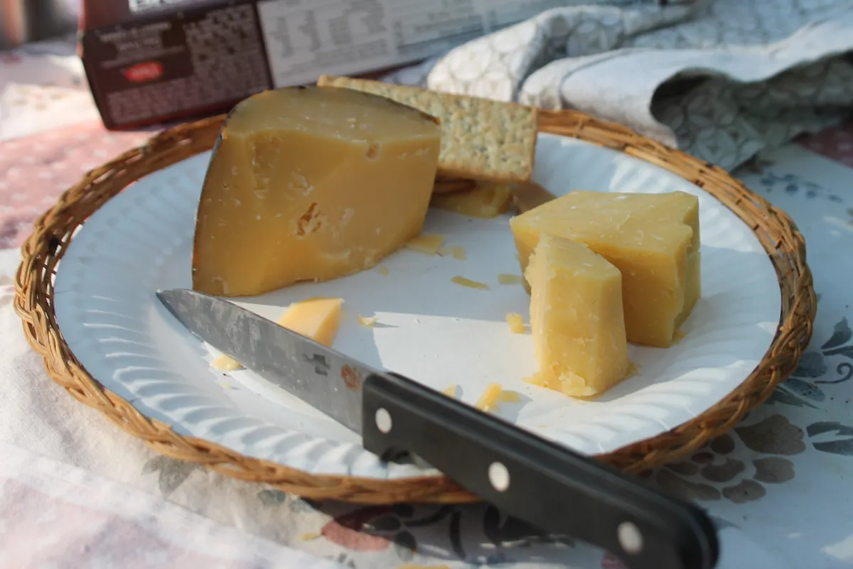 Chunks of aged cheese and crackers on a paper plate in a wicker holder with a knife, set up at a campsite.