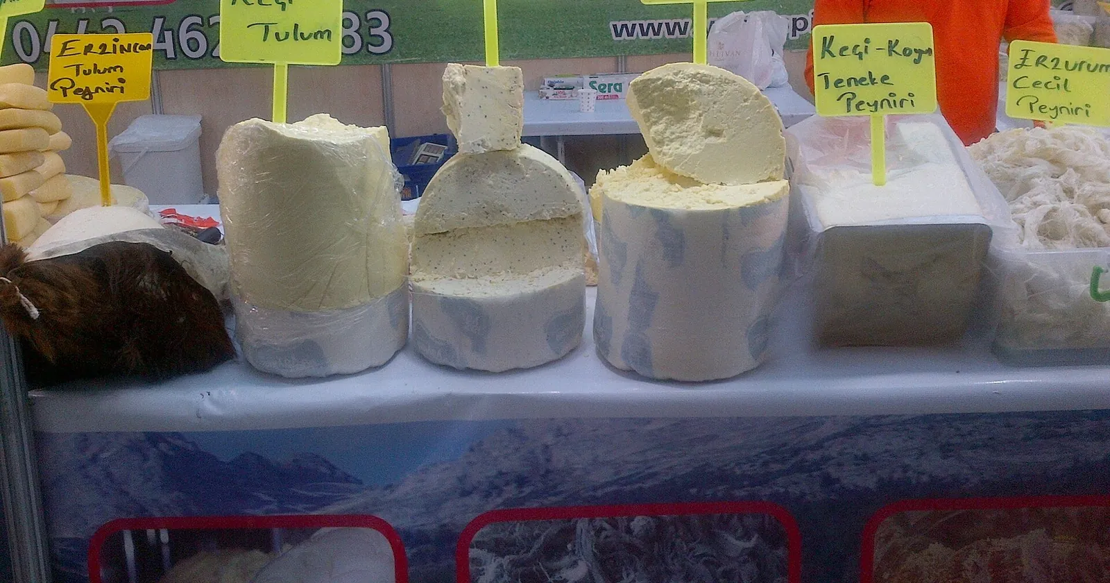 A market stall with white tulum and other Turkish cheeses cut into big pieces, each marked with a yellow label naming the cheese.