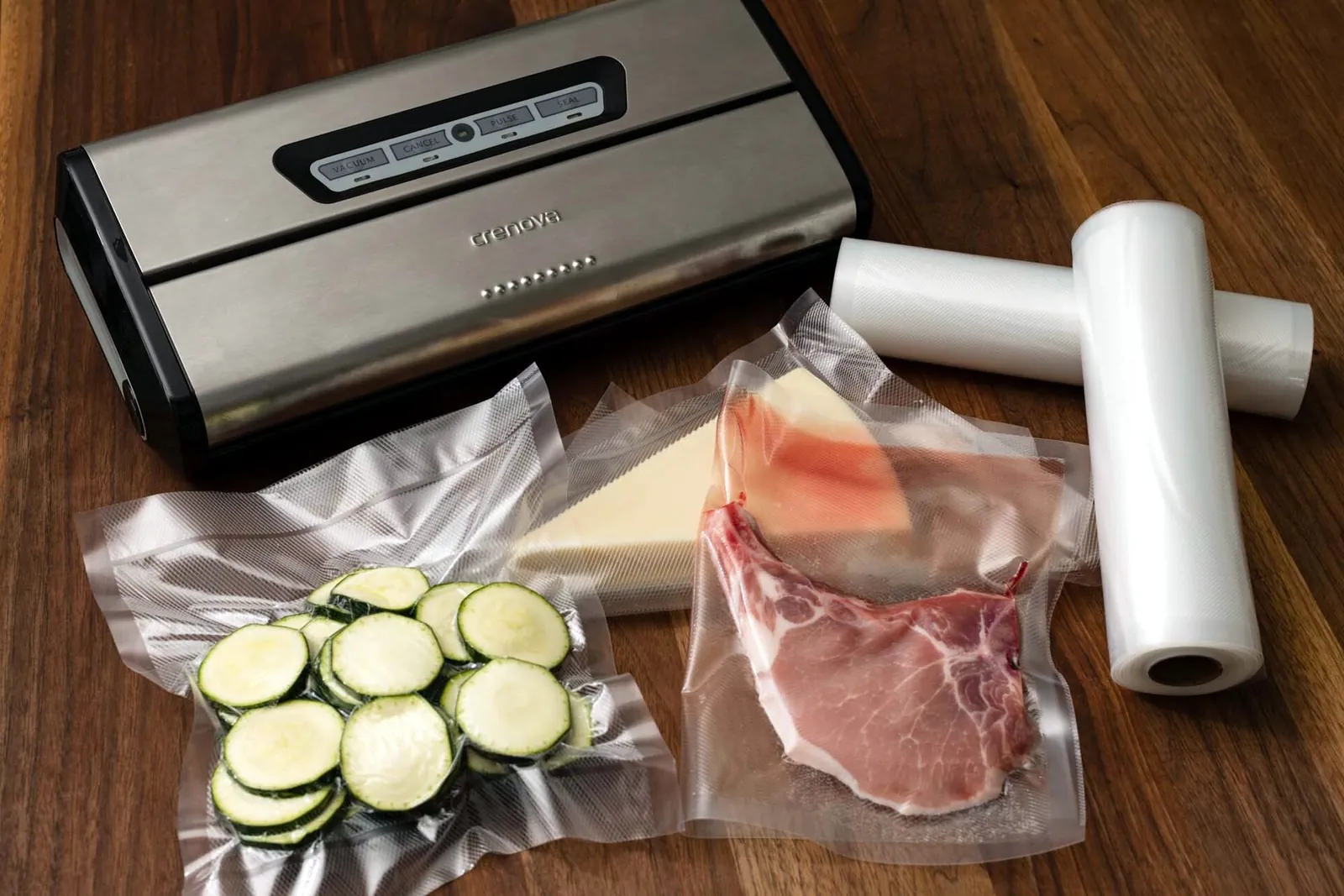 A countertop vacuum sealer with a roll of bag film and sealed bags holding sliced zucchini, a piece of cheese and a pork chop.