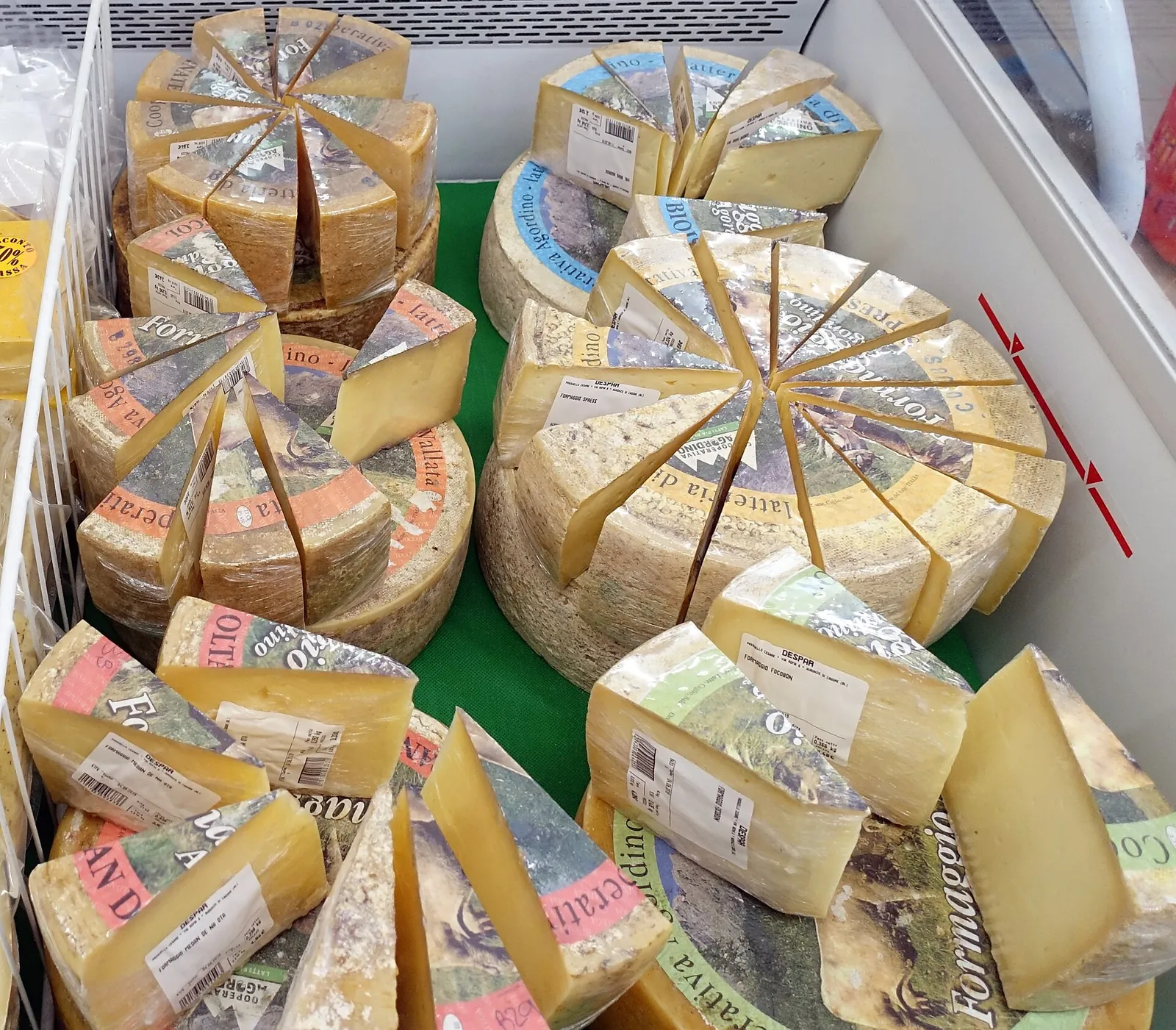 A refrigerated shop case of imported cheeses, with large Italian and French wheels cut into wedges and wrapped pieces arranged in rows