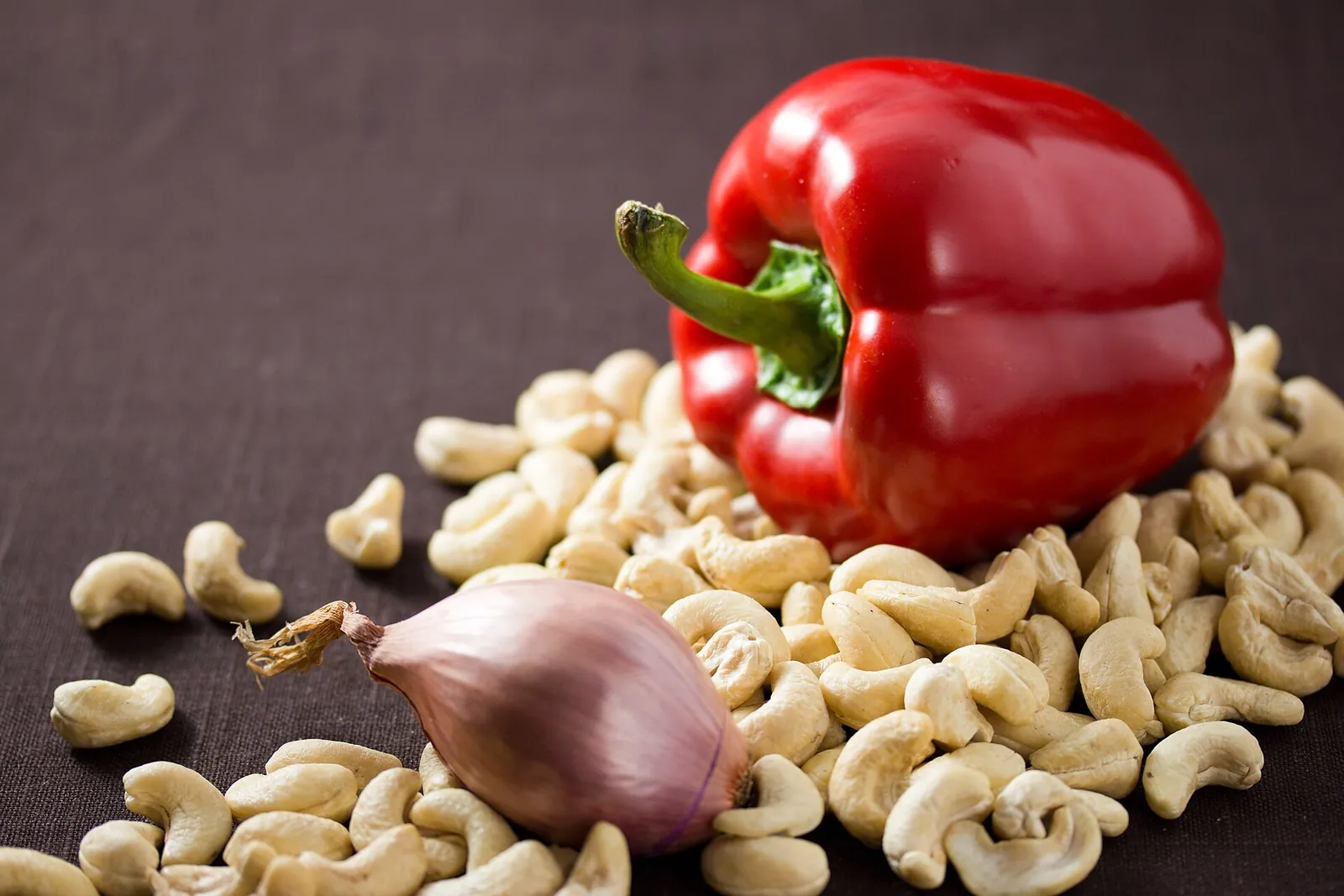 Raw cashew nuts spread on a dark table beside a red bell pepper and a shallot, the base ingredients of cashew cheese.