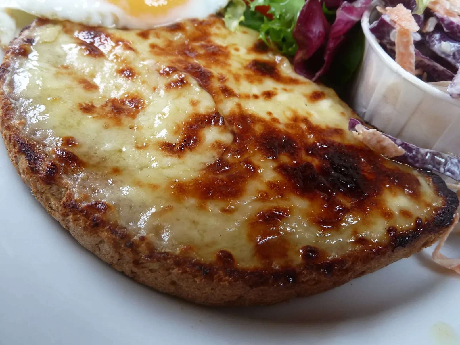 A slice of toast covered in browned, bubbling Welsh rarebit cheese sauce on a white plate with salad