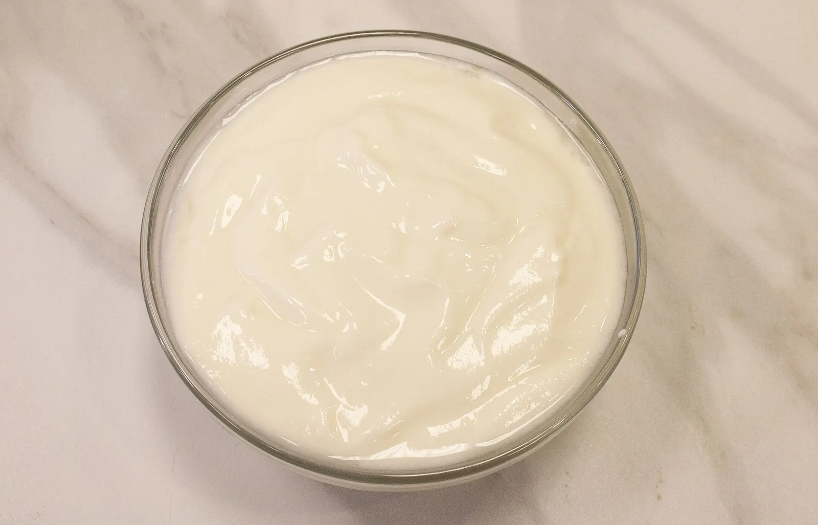 A clear glass bowl of smooth, thick plain yogurt on a pale marble counter, seen from above.